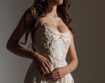 May include: A strapless ivory gown featuring a structured bodice adorned with intricate white floral embroidery, beadwork, and delicate pearl-like swags draped across the chest, transitioning into a full, smooth, floor-length satin skirt.