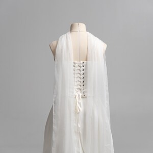 Puede incluir: Un vestido blanco sin mangas con espalda estilo cors&eacute;, ojales de metal plateado y cierre de cordones con cinta blanca. La prenda incluye paneles largos de tul blanco transparente drapeados desde los hombros hacia la espalda.