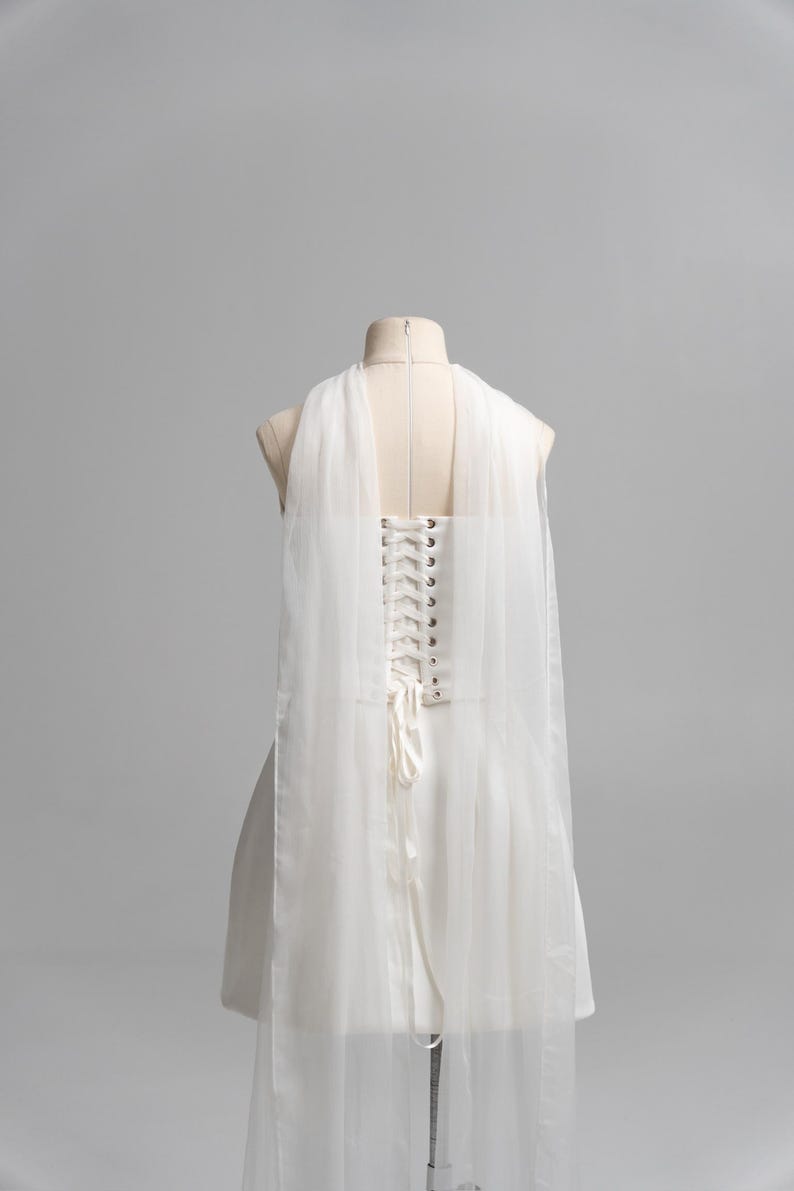Puede incluir: Un vestido blanco sin mangas con espalda estilo cors&eacute;, ojales de metal plateado y cierre de cordones con cinta blanca. La prenda incluye paneles largos de tul blanco transparente drapeados desde los hombros hacia la espalda.
