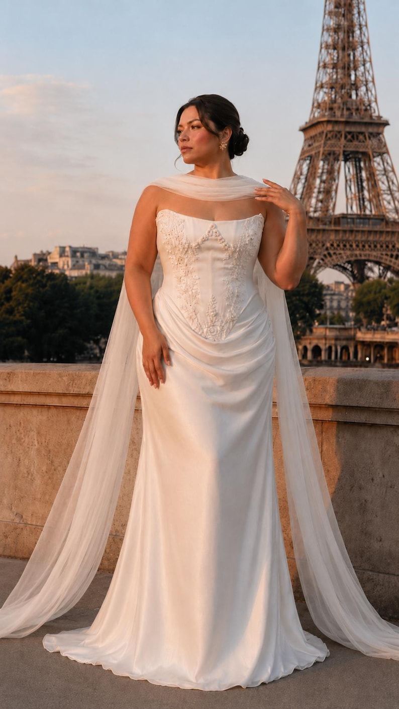 Puede incluir: Un vestido blanco sin tirantes hasta el suelo con un corpi&ntilde;o estructurado que presenta intrincados bordados florales y pedrer&iacute;a. La falda tiene una cintura drapeada y fruncida con una silueta fluida, complementada por una capa larga de tul.