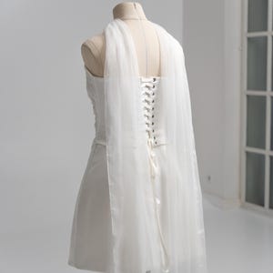 Puede incluir: Un vestido corto blanco sin tirantes sobre un maniqu&iacute;, con espalda estilo cors&eacute;, ojales plateados y cierre de cordones con cinta. El vestido incluye paneles largos de tela blanca transparente y fluida que caen desde los hombros por la espalda.