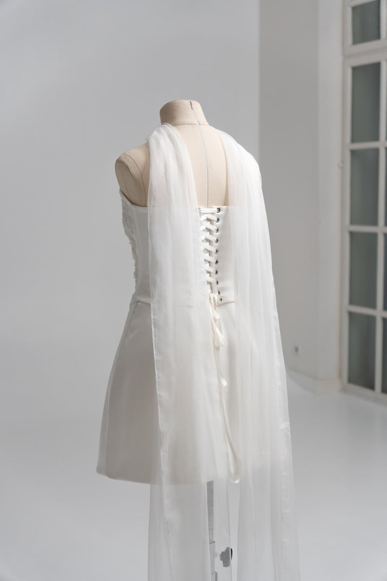Puede incluir: Un vestido corto blanco sin tirantes sobre un maniqu&iacute;, con espalda estilo cors&eacute;, ojales plateados y cierre de cordones con cinta. El vestido incluye paneles largos de tela blanca transparente y fluida que caen desde los hombros por la espalda.