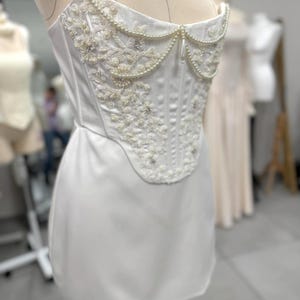 Può includere: Abito a corsetto bianco senza spalline con intricate perline di perle e fiori. Il corpetto presenta cuciture verticali e uno scollo a cuore. L'abito è esposto su un manichino.