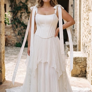 May include: A floor-length white gown featuring a structured corset-style bodice with vertical seams and a square neckline. The dress includes long, wide ribbon straps that tie at the shoulders and a layered, flowing skirt with a textured finish.
