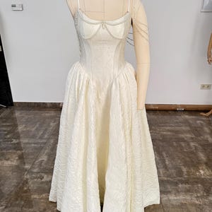 May include: A floor-length cream-colored dress with a textured floral pattern, featuring a fitted bodice, thin spaghetti straps, and a full, pleated skirt. The neckline is adorned with delicate white pearl strands that drape across the chest.