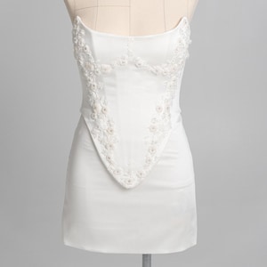 May include: A white corset-style top featuring a straight neckline and a pointed hemline. The front panel is decorated with intricate white floral embroidery and beadwork that follows the bodice seams, creating a structured and detailed appearance.