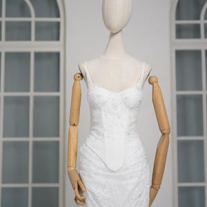 May include: White, embellished bustier dress with spaghetti straps, displayed on a mannequin. The dress features a fitted bodice, a flared skirt, and delicate lace trim. The mannequin has wooden arms and a neutral-colored head.