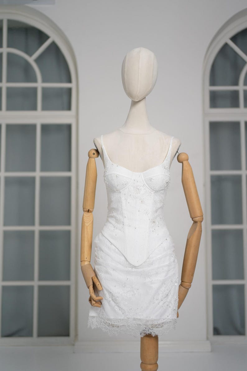 May include: White, embellished bustier dress with spaghetti straps, displayed on a mannequin. The dress features a fitted bodice, a flared skirt, and delicate lace trim. The mannequin has wooden arms and a neutral-colored head.