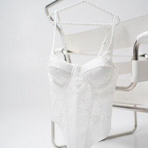 May include: White, embroidered corset top with delicate floral details. The bustier is suspended from a pearl-beaded hanger, showcasing its intricate design. The garment features a structured bodice and thin straps, perfect for special occasions.