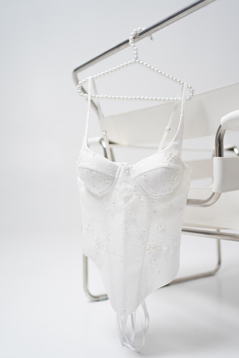 May include: White, embroidered corset top with delicate floral details. The bustier is suspended from a pearl-beaded hanger, showcasing its intricate design. The garment features a structured bodice and thin straps, perfect for special occasions.
