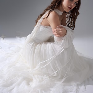 May include: A floor-length white gown featuring a pleated bodice, a high neckline with a collar, and long, sheer, voluminous sleeves. The skirt is constructed from multiple layers of soft, flowing tulle that extend outward across the floor.