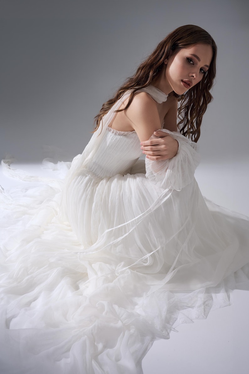 May include: A floor-length white gown featuring a pleated bodice, a high neckline with a collar, and long, sheer, voluminous sleeves. The skirt is constructed from multiple layers of soft, flowing tulle that extend outward across the floor.
