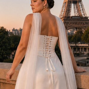 Puede incluir: Un vestido corto blanco sin mangas que presenta una espalda estilo cors&eacute; con detalles de cordones y largos paneles de tela de tul transparente y fluida unidos a los hombros que caen por la parte posterior de la prenda hasta el dobladillo.