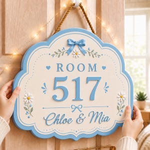 May include: A scalloped, light blue and cream-colored wall sign featuring the text "ROOM 517" and "Chloe & Mia" in blue lettering. The sign is decorated with a raised blue bow, small white daisy flowers, and blue flourishes, suspended by a thick brown rope.