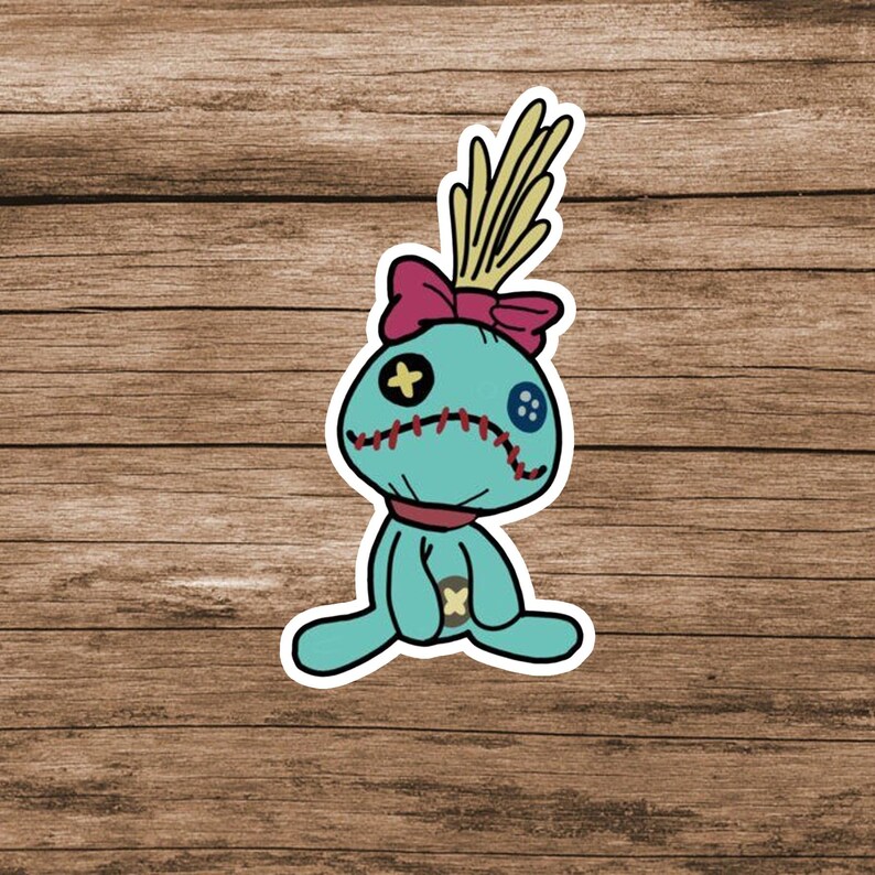 May include: A teal voodoo doll sticker featuring a large pink bow, yellow hair, and a stitched mouth. The doll has one yellow cross-shaped eye and one blue button eye, with a small yellow cross on its torso, all outlined in white.