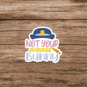 May include: A die-cut sticker featuring a dark blue police hat with a yellow badge on top. Below the hat, the text reads NOT YOUR AVERAGE BUNNY in pink, yellow, and light blue letters. An orange carrot graphic is positioned next to the word AVERAGE.