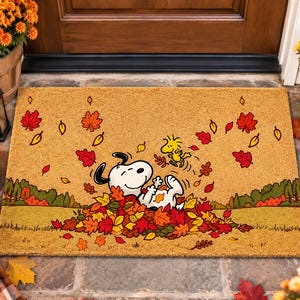 May include: A rectangular tan coir doormat featuring a printed illustration of Snoopy and Woodstock playing in a pile of colorful autumn leaves. The scene includes red, orange, and yellow foliage scattered across a grassy field background.