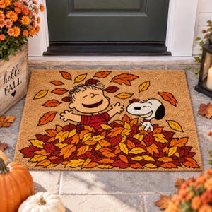 May include: A rectangular tan coir doormat featuring a printed illustration of the Peanuts characters Charlie Brown and Snoopy playing in a large pile of colorful autumn leaves, with several leaves floating in the air above them.