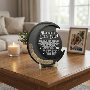May include: A black, crescent-shaped coin holder with the text "Heaven's Little Coin" and a poem. The holder is propped up on a white stand and filled with coins. A framed family photo and a lit candle are in the background.