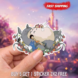May include: A die-cut sticker featuring a large grey Totoro character with a beige belly, a smaller blue Totoro, and a human figure resting on the grey creature. The design includes small white spirits, dark soot sprites, and colorful floral accents.