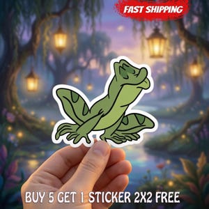 May include: A die-cut sticker featuring a cartoon green frog with dark green spots, shown in a profile view with its head tilted upward. The sticker has a white border and is set against a blurred background of a forest with hanging lanterns.
