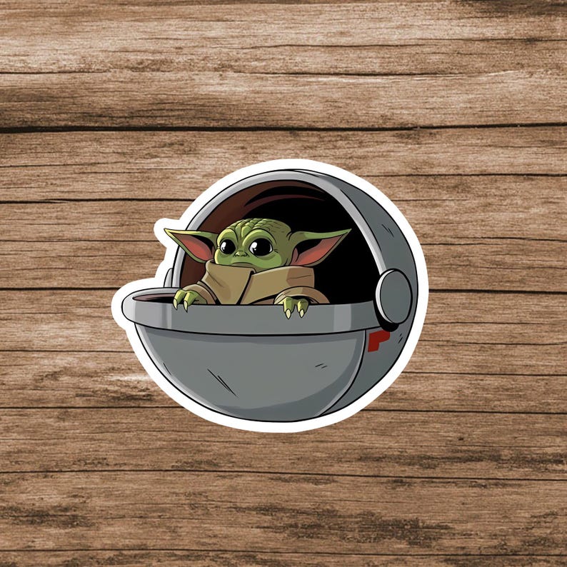 May include: A die-cut sticker featuring a green, large-eared creature with big black eyes, wearing a tan robe, sitting inside a round, metallic gray hover pod with a dark interior and a small red accent mark on the side of the exterior shell.