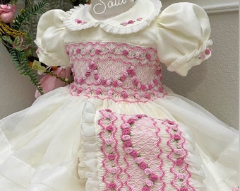 Peut inclure: Une robe blanche smockée avec des broderies florales roses et un coussin assorti. La robe a un col à volants et des manches bouffantes. La robe est ornée d'un bandeau blanc avec un nœud rose.