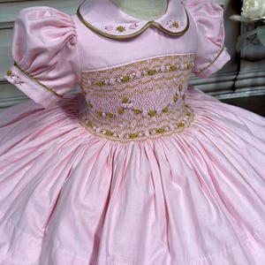 以下が含まれることがあります： 白い襟と金のトリムが付いたピンクのフリルドレス。ドレスは短いパフスリーブとフルスカート。ピンクとゴールドの刺繍の花で飾られています。