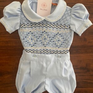 以下が含まれることがあります： 水色のベビー服セット。スモック加工のボディスと、お揃いのブルマが特徴です。ピーターパンカラーとパフスリーブ、白い刺繍が施されています。「Solu's Designs」と書かれたタグが付いています。