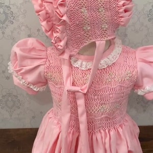 May include: A pink smocked baby dress with a matching bonnet. The dress has a white lace trim and a pink ribbon tied at the back. The bonnet has a ruffled edge. The dress is embroidered with a floral pattern.