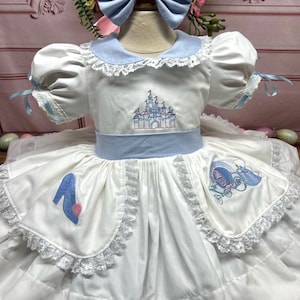 以下が含まれることがあります： 青いアクセントが付いた白いドレスで、城とガラスの靴の刺繍が施されています。ドレスはレースのトリムと青いリボンヘッドバンドが付いています。