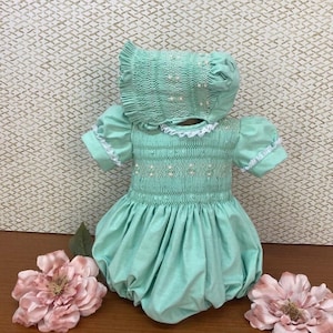 May include: A mint green smocked baby dress with a matching bonnet. The dress has a white lace trim and pearl-like embellishments.