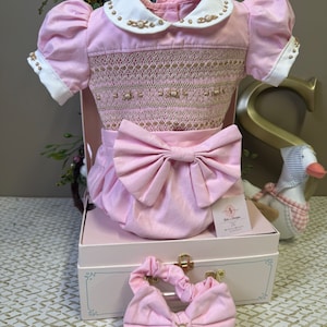 以下が含まれることがあります： 白い襟と金の刺繍が施されたピンクのベビー服。ドレスは正面に大きなピンクのリボンが付いており、ピンクのギフトボックスにピンクのリボンを合わせた状態で展示されています。