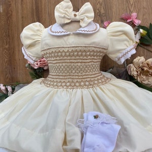 以下が含まれることがあります： 白いスモックドレスに、茶色の刺繍模様が施されています。白い襟と白いリボンが付いています。ドレスはパフスリーブで、スカートはボリュームがあります。レースのトリムと小さな花のデザインが施された白い靴下1足が付属しています。