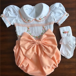 May include: A white smocked baby outfit with a peach-colored bow and matching bloomers. The outfit is embroidered with small pink and green flowers. The outfit is labeled "12M".