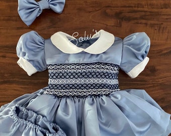 Peut inclure: Une robe en satin bleu clair avec un col blanc et des manches bouffantes. La robe a un corsage smocké et une jupe ample. Elle est accompagnée d'un bandeau en satin bleu clair assorti avec un nœud. La robe est sur une surface en bois.