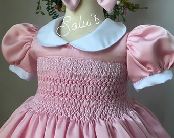 Peut inclure: Une robe rose smockée avec un col blanc Peter Pan et des manches bouffantes. La robe a une finition satinée et est ornée d'un nœud rose.