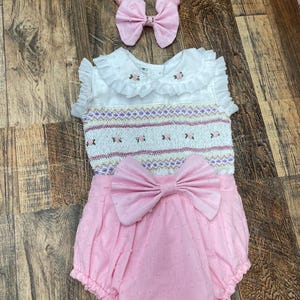 以下が含まれることがあります： 白とピンクのベビー服。白い刺繍のトップス、ピンクのドット柄のブルマ、ピンクのリボン付きヘッドバンドです。