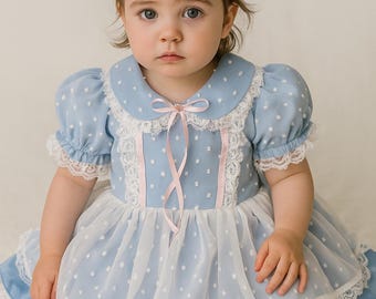 Puede incluir: Una bebé con un vestido azul claro con lunares blancos, ribete de encaje blanco y un lazo rosa. El vestido tiene cuello Peter Pan y mangas abullonadas. Una diadema con lazo rosa adorna la cabeza del bebé. El texto "2 años" es visible.