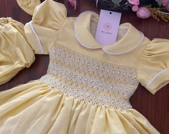May include: A yellow smocked dress with white trim and a white collar. The dress has puffed sleeves and a full skirt. The dress is made of a soft, lightweight fabric.