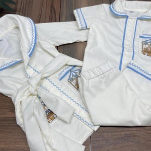 以下が含まれることがあります： 子供用のパジャマと、お揃いのバスローブのセット。白い衣服には、水色のトリムと刺繍のディテールが施されています。パジャマのトップスには、ポケットにメリーゴーランドのデザインがあります。バスローブにはお揃いのベルトが付いています。