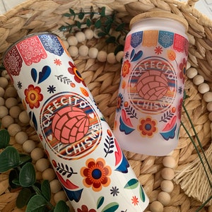 May include: Two white tumblers with a colorful floral design and the text "Cafecito y Chismes". The tumblers are sitting in a woven basket with wooden beads.