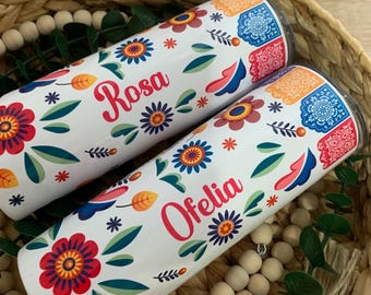 Op de afbeelding: Twee witte bekers met kleurrijke bloemenpatronen en de namen "Rosa" en "Ofelia" erop gedrukt. De bekers staan in een gevlochten mand met houten kralen.