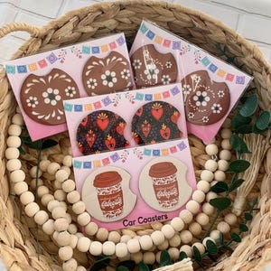 May include: Set of six colorful car coasters featuring various designs. The coasters are packaged in clear plastic and include designs with flowers, hearts, and coffee cups. The text on the coffee cup coasters reads "wonderful sweet cafécito".