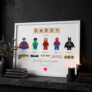 Könnte beinhalten: Ein gerahmter Druck mit dem Wort DADDY aus Holzbuchstaben, gefolgt von fünf Superhelden-Minifiguren, beschriftet als Fast As Superman, Strong As Hulk, Smart As Iron Man, Funny As Spider-Man und Brave As Batman, mit dem Text You're My Favourite Superhero! Love Freya x und einem roten Herz.
