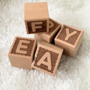 Puede incluir: Bloques de alfabeto de madera con letras grabadas. Los bloques son de color marrón claro con letras y bordes decorativos de color marrón oscuro. Las letras visibles son F, E, A e Y. Los bloques están dispuestos sobre una superficie blanca y esponjosa.