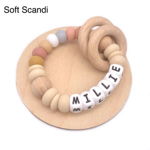 May include: A wooden teething ring with a silicone bead bracelet featuring the name "Millie" in white beads with black lettering. The beads are in various colors, including beige, light brown, mustard yellow, and light pink.