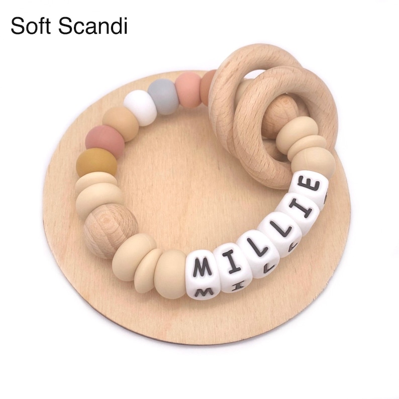May include: A wooden teething ring with a silicone bead bracelet featuring the name "Millie" in white beads with black lettering. The beads are in various colors, including beige, light brown, mustard yellow, and light pink.