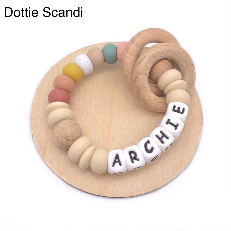 May include: A wooden teething ring with a colorful silicone bead bracelet and a white silicone bead necklace with the name "ARCHIE" spelled out.