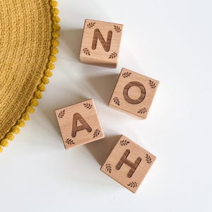 Puede incluir: Cuatro bloques de letras de madera que deletrean "N O A H". Cada bloque es de color marrón claro con una letra tallada y un diseño de hojas. Los bloques están dispuestos sobre una superficie blanca, con un objeto circular amarillo y texturizado en el fondo.